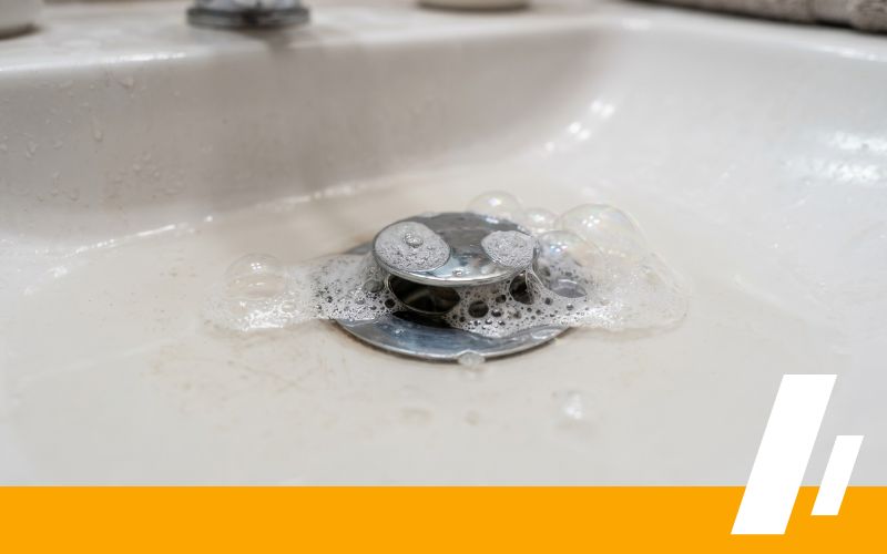 Drain Cleaning 2 - Dirt Cheap Sewer Co. Coins piled on a sink drain with soap bubbles around them, suggesting a clogged drain