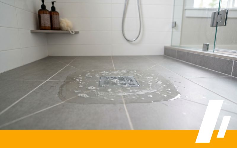 Shower floor with a circular wet patch around the drain, gray tiles, glass door, and a shelf with bottles in the corner.