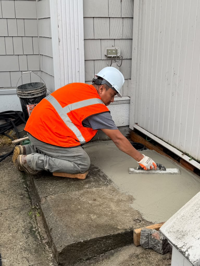 concrete-replacement-water-line - Dirt Cheap Sewer Co. Construction worker in orange safety vest and white helmet smoothing wet concrete with a trowel on a small exterior landing by the building shortly after repairing a water line.