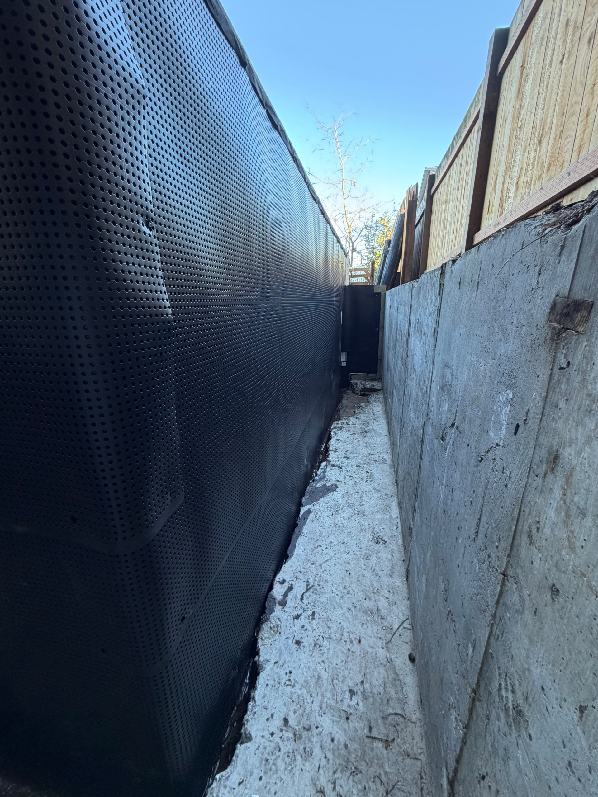 Narrow alley between a tall black perforated metal wall on the left and a wooden fence on the right, implementing foundation drainage on a property
